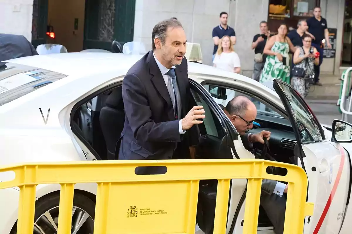 Un hombre con traje sale de un taxi blanco mientras otra persona permanece en el asiento del conductor y varias personas observan desde la acera detrás de una valla amarilla