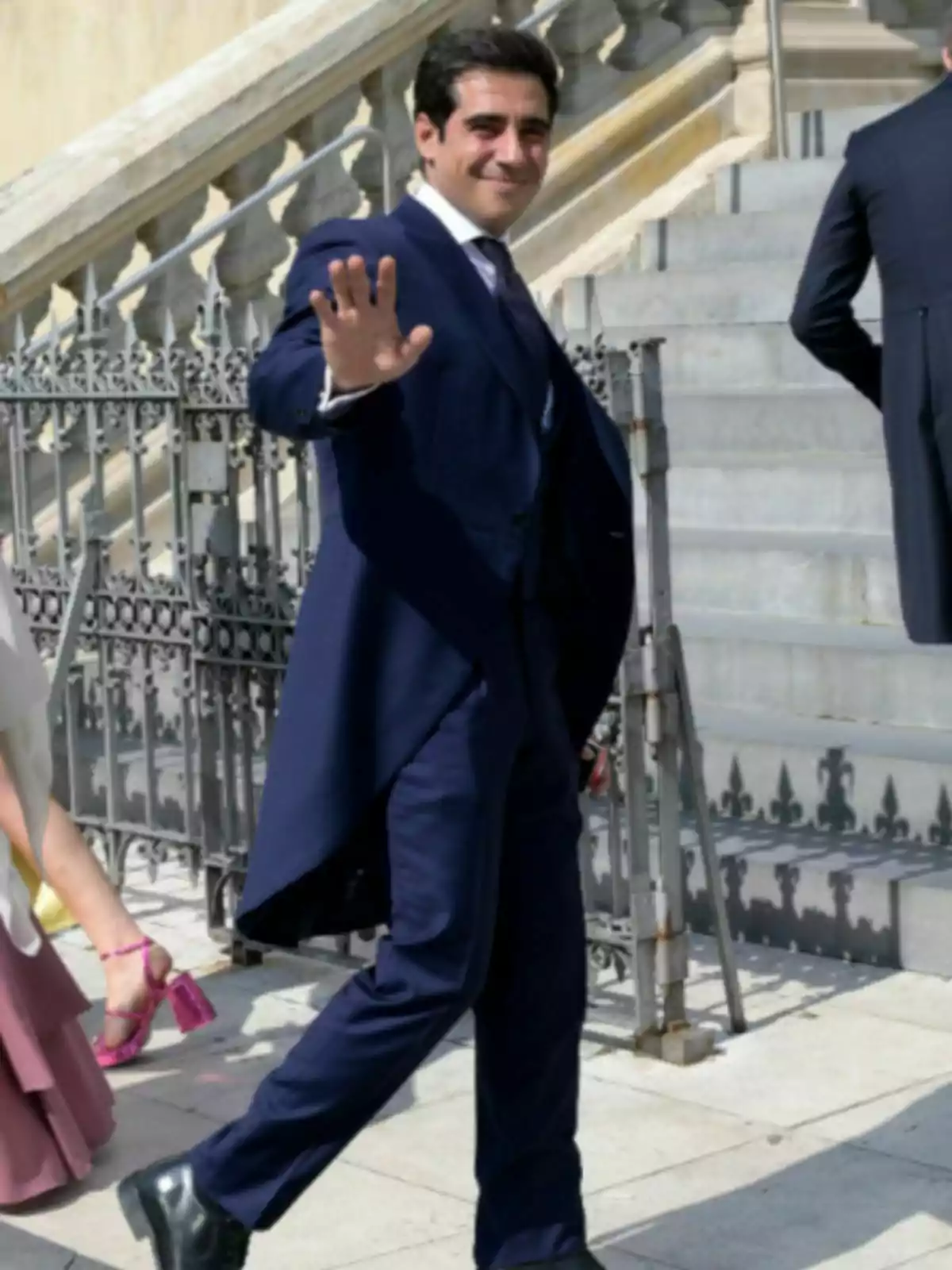 Alberto Herrera con traje azul oscuro saludando y sonriendo mientras camina por la calle frente a unas escaleras de piedra Alberto Herrera con traje azul oscuro saludando y sonriendo mientras camina por la calle frente a unas escaleras de piedra
