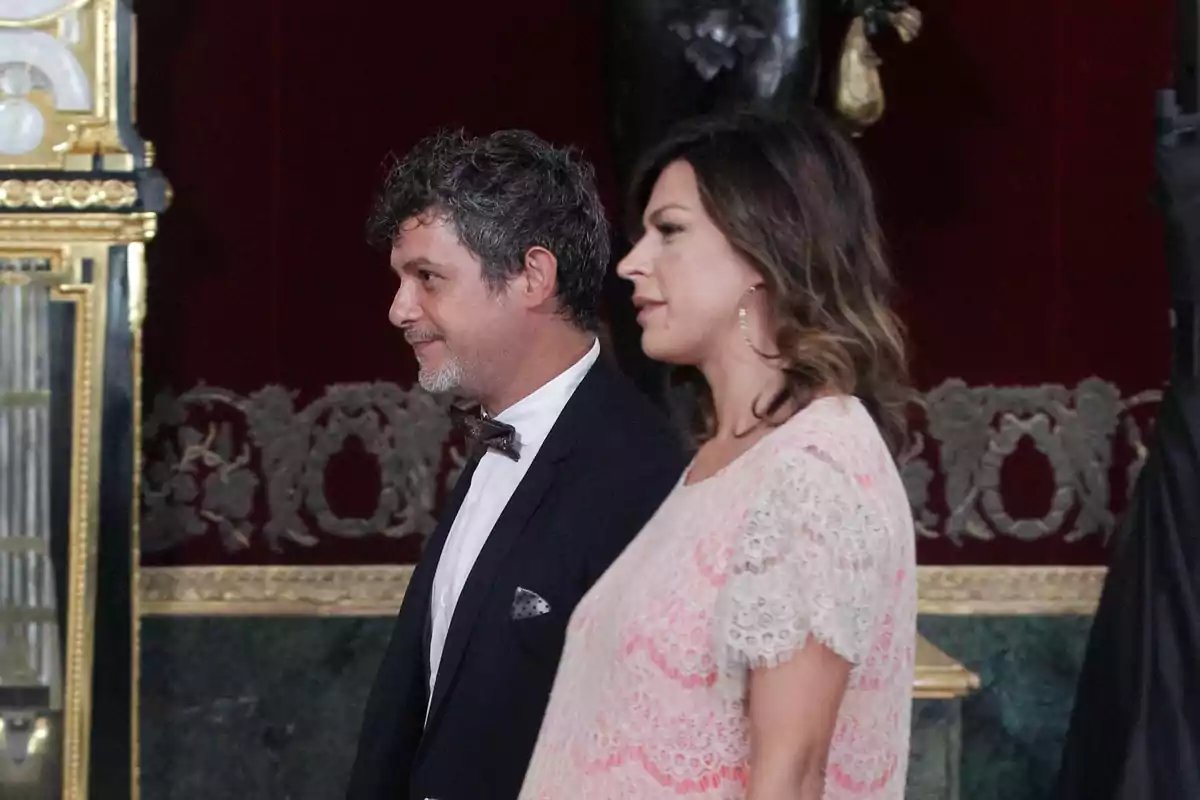 Alejandro Sanz in a tuxedo and Raquel Perera in a light-colored dress walk together in an elegant hall decorated with golden details and red walls. Alejandro Sanz in a tuxedo and Raquel Perera in a light-colored dress walk together in an elegant hall decorated with golden details and red walls.