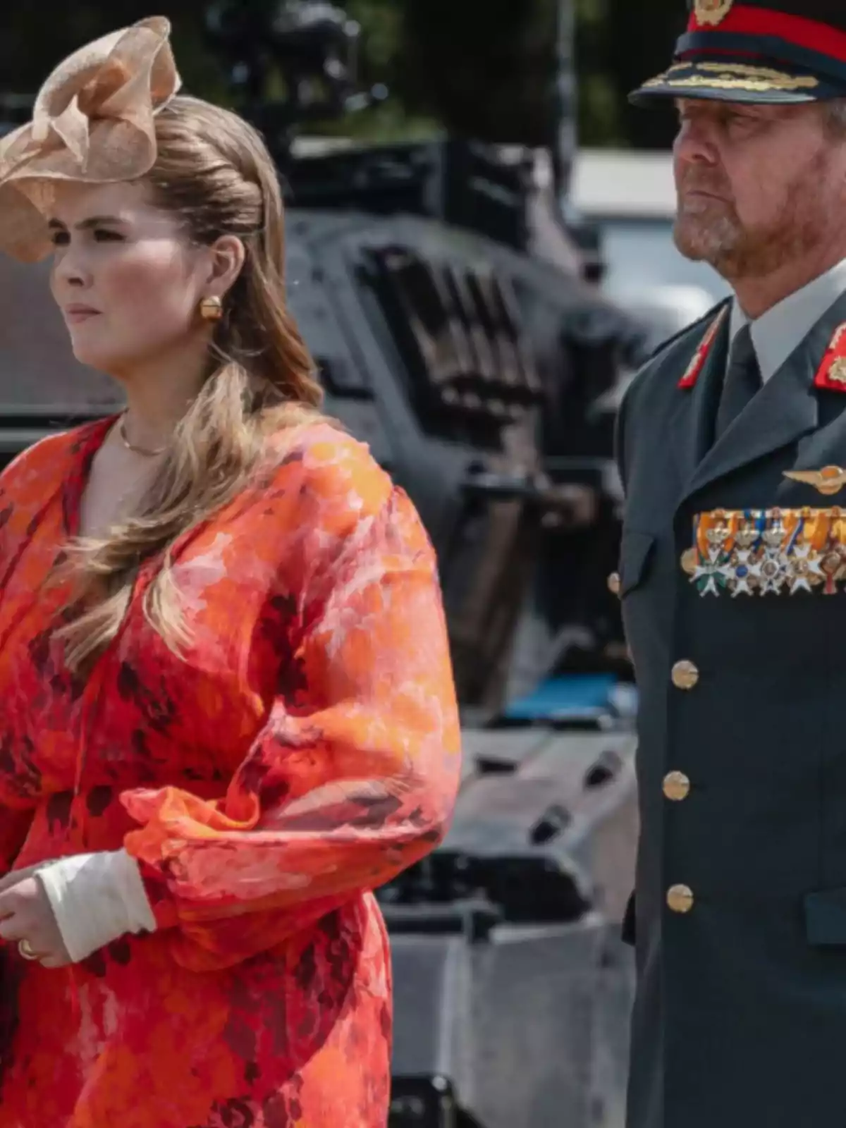 A woman in a red dress and an elegant headpiece stands next to a man in a military uniform with medals, both standing in front of an armored vehicle.