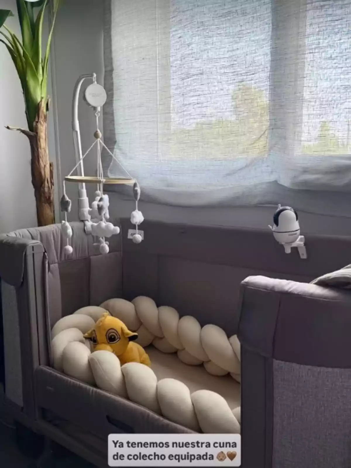 Publicación de Asraf Beno en Instagram donde se ve la cuna de colecho gris para su bebé, que está situada junto a una ventana con cortinas claras, decorada con un móvil de peluches y un cojín trenzado beige Publicación de Asraf Beno en Instagram donde se ve la cuna de colecho gris para su bebé, que está situada junto a una ventana con cortinas claras, decorada con un móvil de peluches y un cojín trenzado beige