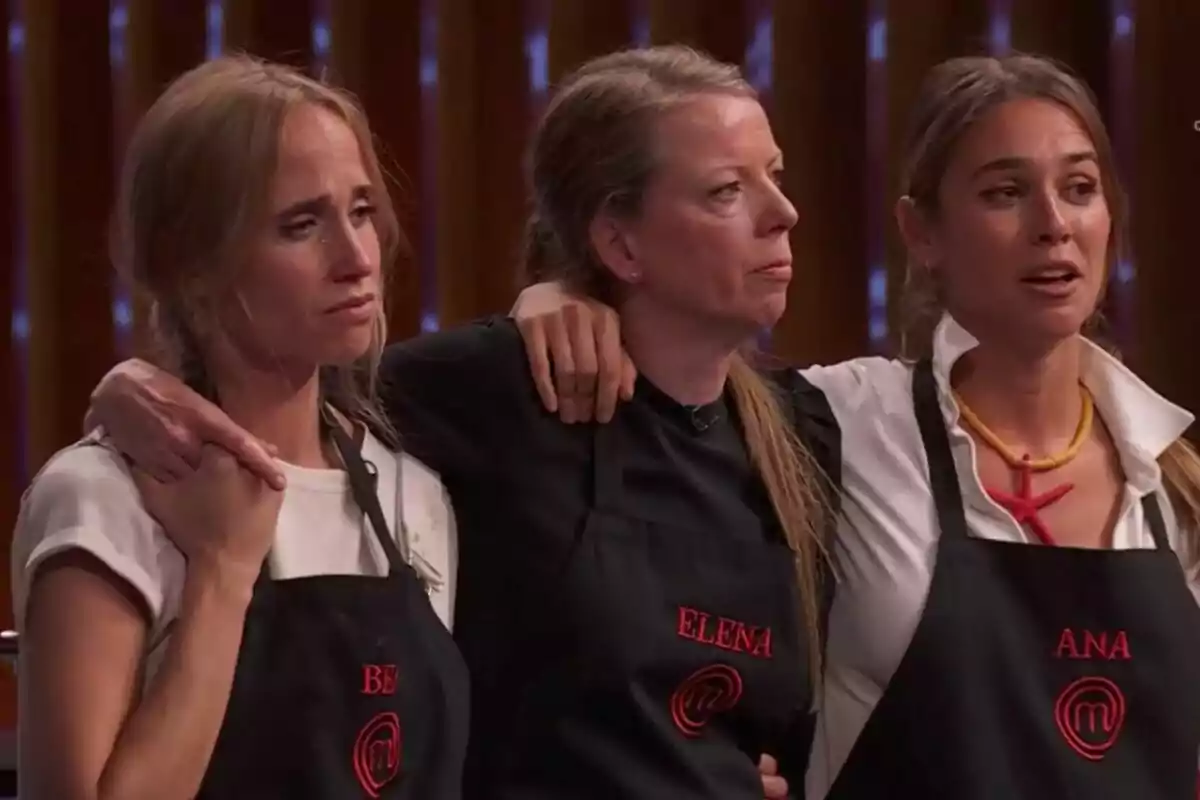 Tres mujeres -Bea, Elena y Ana- con delantales negros de MasterChef se abrazan y muestran expresiones de emoción y nerviosismo en un set de cocina iluminado. Tres mujeres -Bea, Elena y Ana- con delantales negros de MasterChef se abrazan y muestran expresiones de emoción y nerviosismo en un set de cocina iluminado.