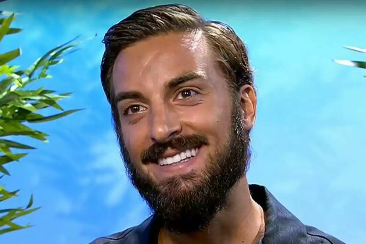 Borja González joven con barba y cabello corto sonríe frente a un fondo azul con plantas a los lados en Supervivientse Borja González joven con barba y cabello corto sonríe frente a un fondo azul con plantas a los lados en Supervivientse