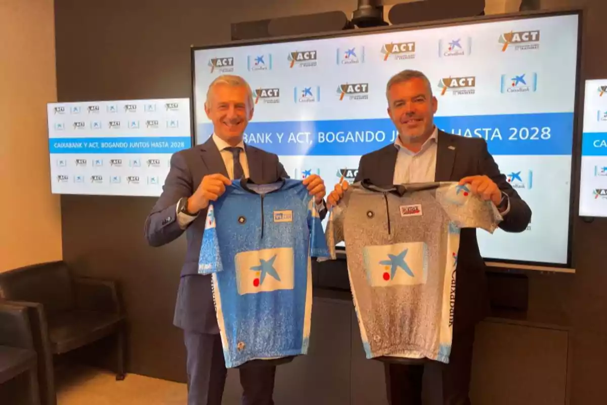 Dos hombres de traje sonríen mientras sostienen camisetas deportivas frente a una pantalla con logotipos de Caixabank y ACT Dos hombres de traje sonríen mientras sostienen camisetas deportivas frente a una pantalla con logotipos de Caixabank y ACT