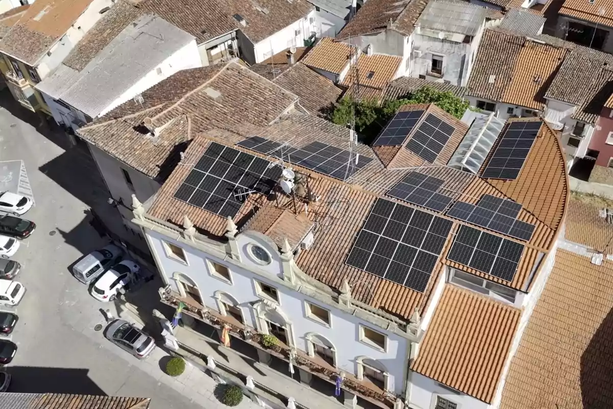 Vista aérea de un edificio con tejado de tejas naranjas cubierto con paneles solares rodeado de otros edificios y coches estacionados en la calle Vista aérea de un edificio con tejado de tejas naranjas cubierto con paneles solares rodeado de otros edificios y coches estacionados en la calle