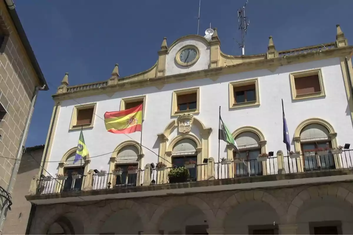 Fachada de un edificio histórico con varias banderas ondeando en el balcón y un reloj en la parte superior