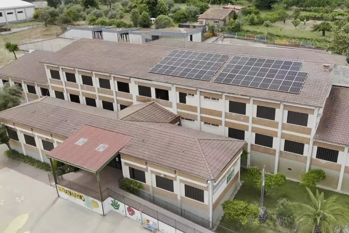 Vista aérea de un edificio escolar de dos pisos con paneles solares en el techo rodeado de vegetación y un patio delantero Vista aérea de un edificio escolar de dos pisos con paneles solares en el techo rodeado de vegetación y un patio delantero