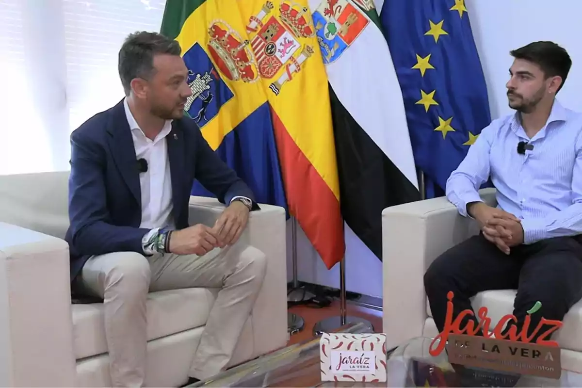 Dos hombres sentados conversando en una oficina con varias banderas oficiales de fondo y un letrero que dice Jaraíz de la Vera sobre la mesa de cristal Dos hombres sentados conversando en una oficina con varias banderas oficiales de fondo y un letrero que dice Jaraíz de la Vera sobre la mesa de cristal
