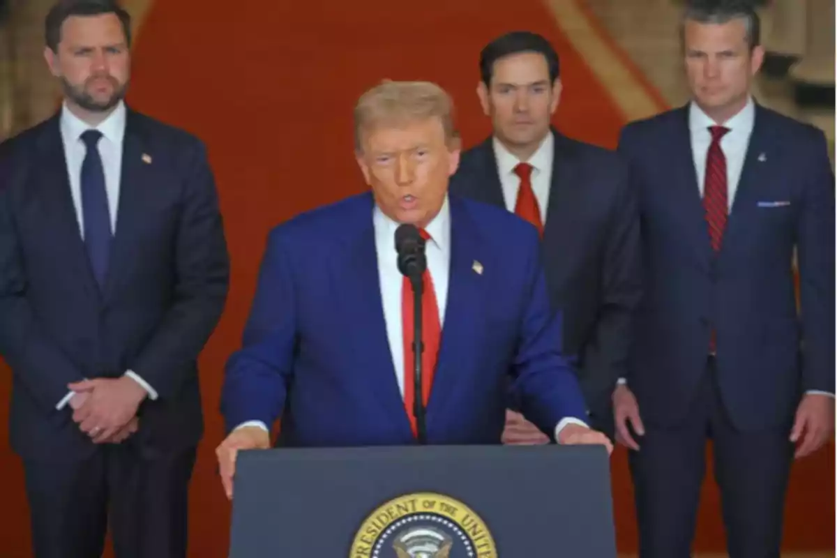 Un hombre de traje azul y corbata roja habla en un podio con el sello presidencial de Estados Unidos mientras tres hombres de traje lo acompañan al fondo Un hombre de traje azul y corbata roja habla en un podio con el sello presidencial de Estados Unidos mientras tres hombres de traje lo acompañan al fondo