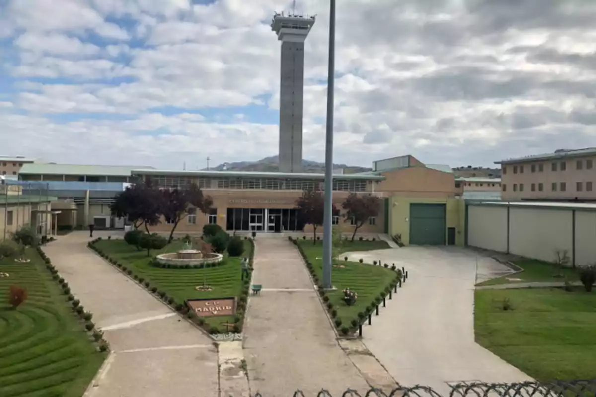 Edificio de una prisión con jardines bien cuidados al frente y una torre de vigilancia alta en el centro bajo un cielo parcialmente nublado Edificio de una prisión con jardines bien cuidados al frente y una torre de vigilancia alta en el centro bajo un cielo parcialmente nublado