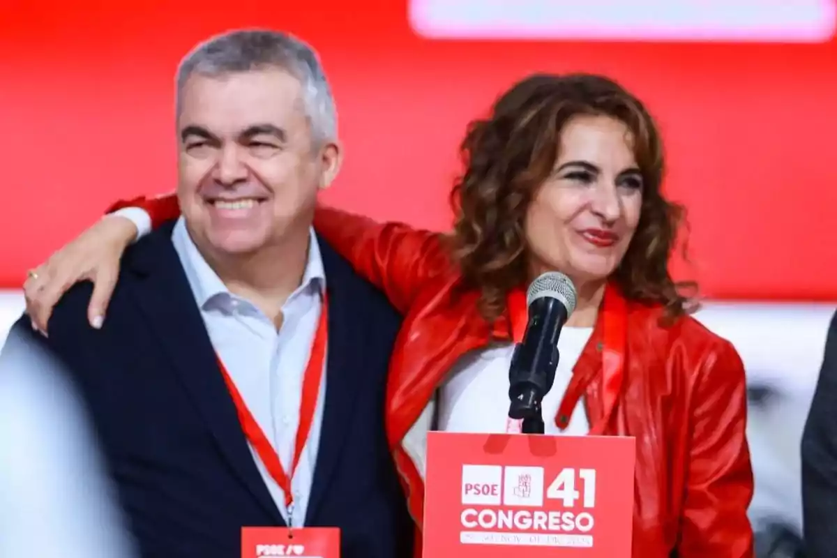Dos personas sonríen abrazadas frente a un micrófono en un evento del PSOE con fondo rojo y cartel del 41 Congreso Dos personas sonríen abrazadas frente a un micrófono en un evento del PSOE con fondo rojo y cartel del 41 Congreso