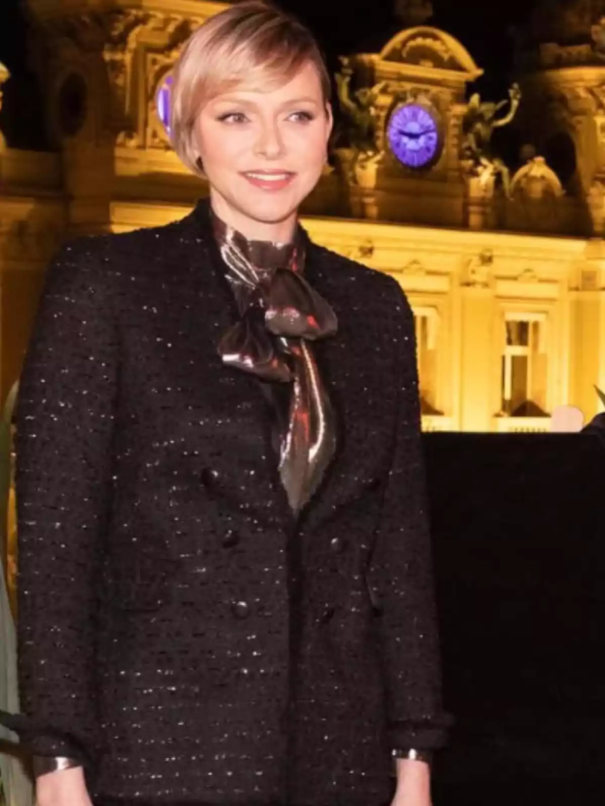Blonde woman with short hair wearing a black blazer and a blouse with a metallic bow, posing in front of a brightly lit building at night with a blue clock on the facade. Blonde woman with short hair wearing a black blazer and a blouse with a metallic bow, posing in front of a brightly lit building at night with a blue clock on the facade.