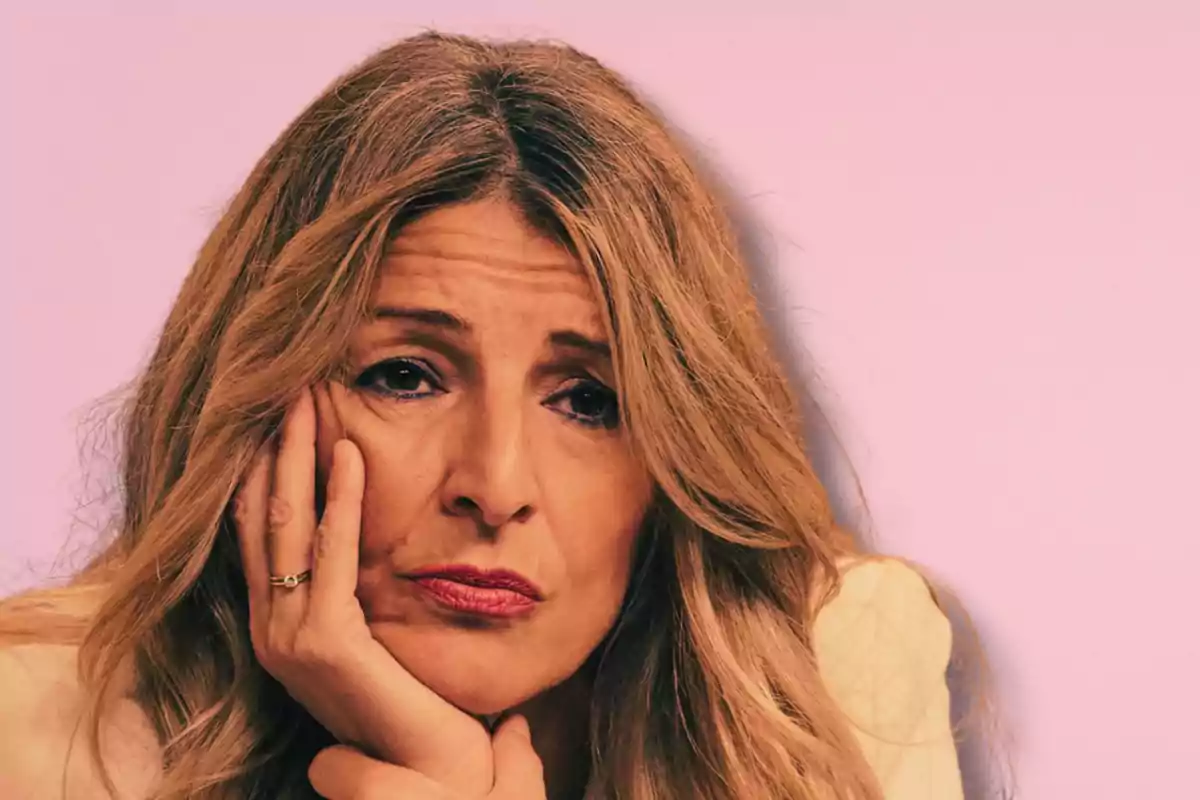 Mujer de cabello largo y rubio con expresión pensativa apoyando la cara en su mano frente a un fondo rosa