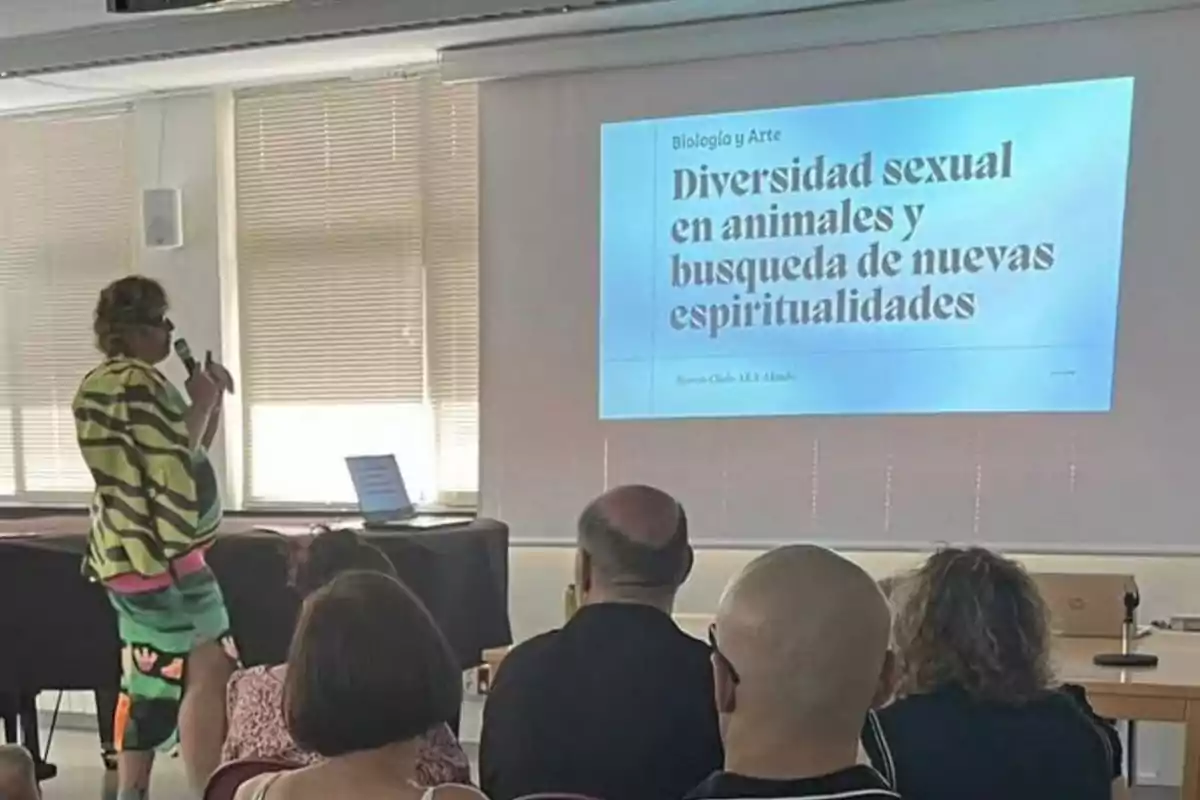 Persona con ropa colorida dando una presentación sobre diversidad sexual en animales y búsqueda de nuevas espiritualidades frente a un grupo de personas en un aula.
