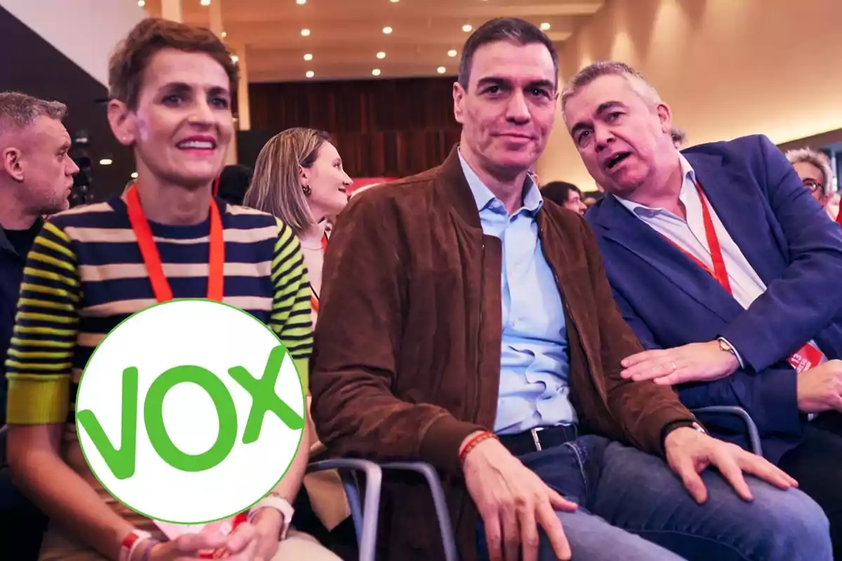 Tres personas sentadas en un evento con acreditaciones rojas y un logo de VOX superpuesto sobre la persona de la izquierda