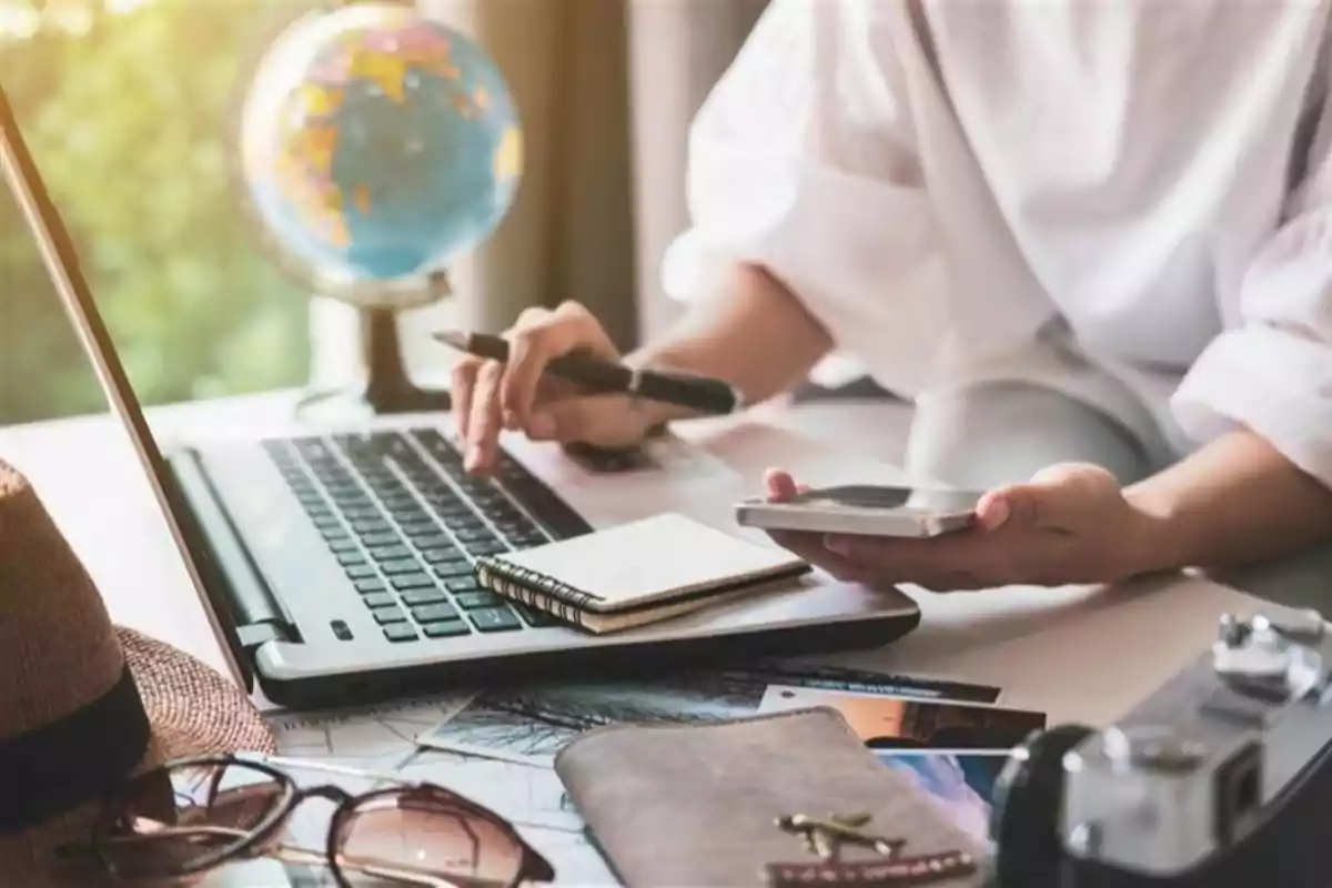 Persona usando una laptop mientras sostiene un teléfono móvil, con una libreta, pasaporte, cámara, gafas de sol y un globo terráqueo sobre la mesa, en un ambiente de planificación de viaje.