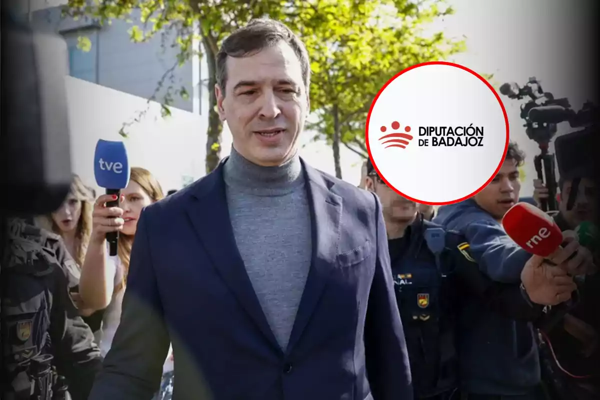 Hombre con chaqueta azul y suéter gris rodeado de periodistas y cámaras al aire libre con un logo de la Diputación de Badajoz en la imagen