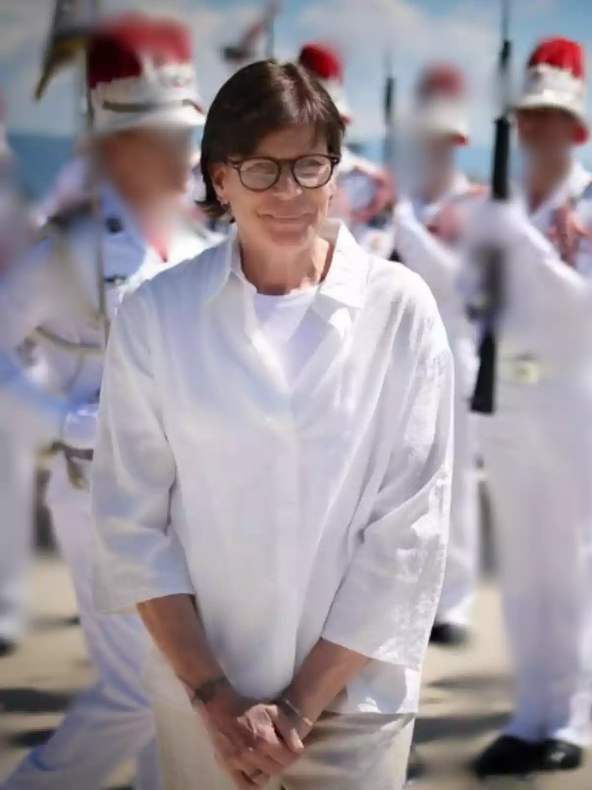 Short-haired woman with glasses dressed in white smiling outdoors with uniformed, blurred people in the background Short-haired woman with glasses dressed in white smiling outdoors with uniformed, blurred people in the background