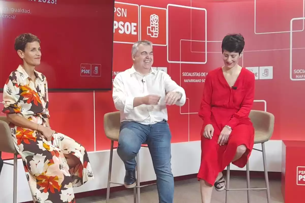 Tres personas sentadas en sillas altas conversan y sonríen en un set con fondo rojo decorado con logotipos del PSOE Tres personas sentadas en sillas altas conversan y sonríen en un set con fondo rojo decorado con logotipos del PSOE