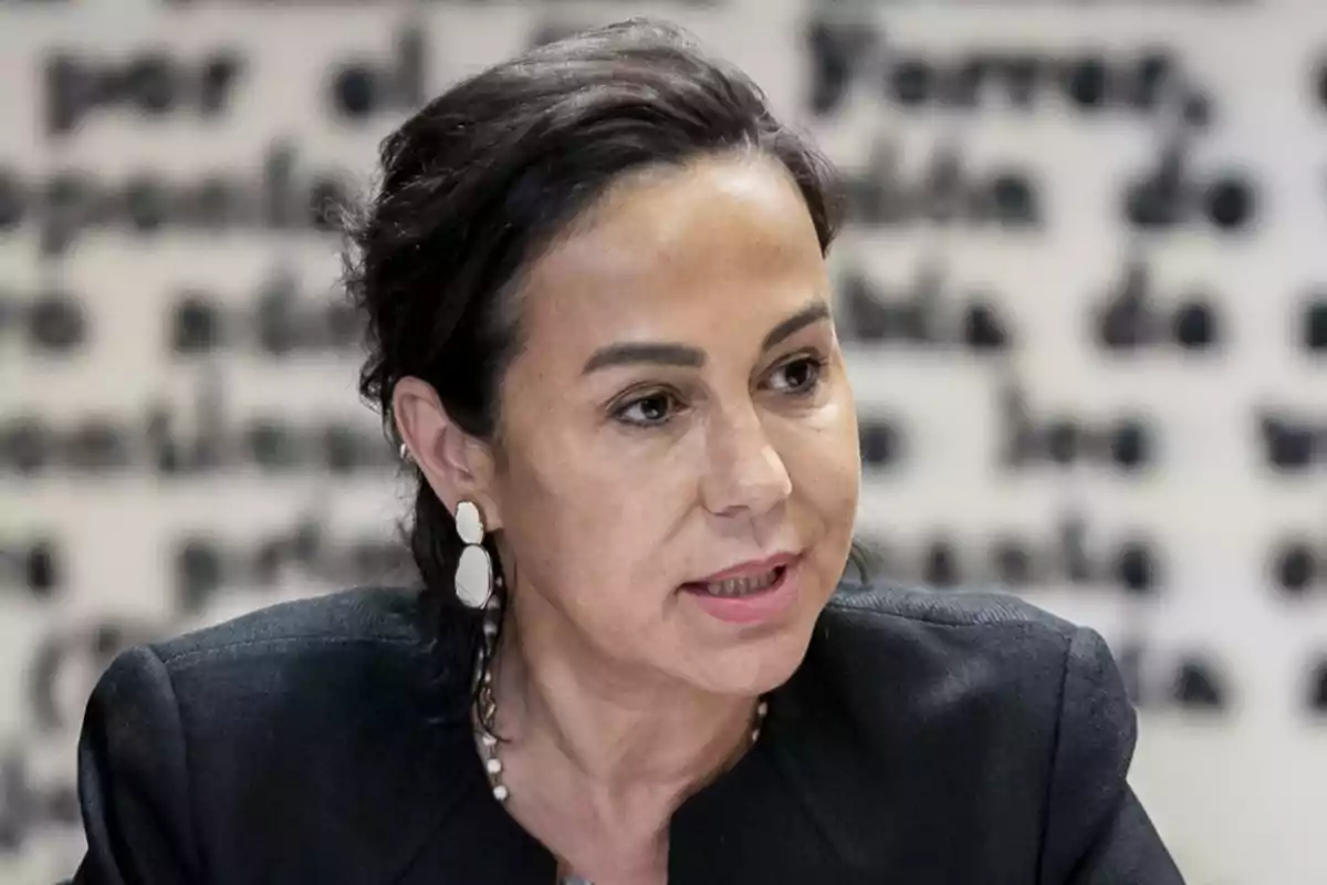 Mujer de cabello oscuro con aretes grandes y chaqueta negra hablando frente a un fondo borroso con letras negras Mujer de cabello oscuro con aretes grandes y chaqueta negra hablando frente a un fondo borroso con letras negras