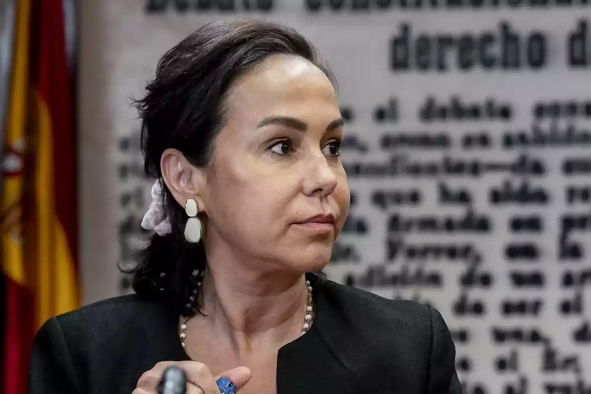 Mujer de cabello oscuro recogido con una liga clara y pendientes grandes, vestida de manera formal, mirando hacia un lado con expresión seria, fondo con texto borroso y una bandera de España a la izquierda