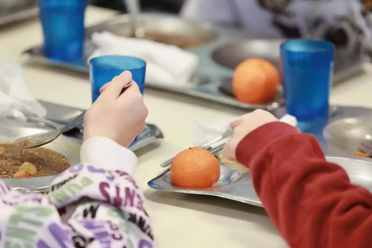 Dos niños comen en una mesa con bandejas de comida, vasos azules y mandarinas