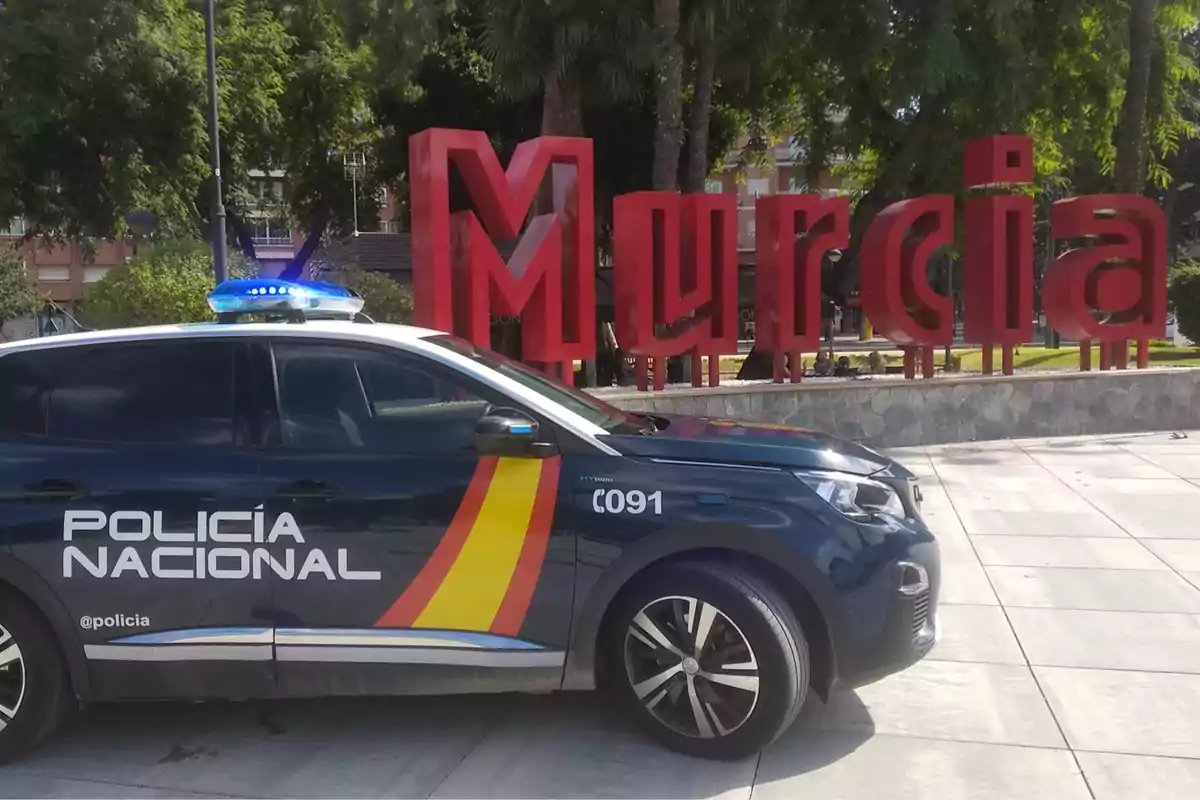 Coche de la Policía Nacional estacionado frente a un letrero grande y rojo que dice Murcia en una zona urbana con árboles al fondo Coche de la Policía Nacional estacionado frente a un letrero grande y rojo que dice Murcia en una zona urbana con árboles al fondo