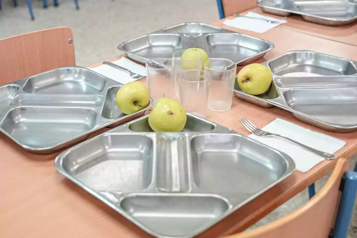 Bandejas metálicas vacías con manzanas verdes, vasos de agua y cubiertos sobre una mesa en un comedor escolar Bandejas metálicas vacías con manzanas verdes, vasos de agua y cubiertos sobre una mesa en un comedor escolar