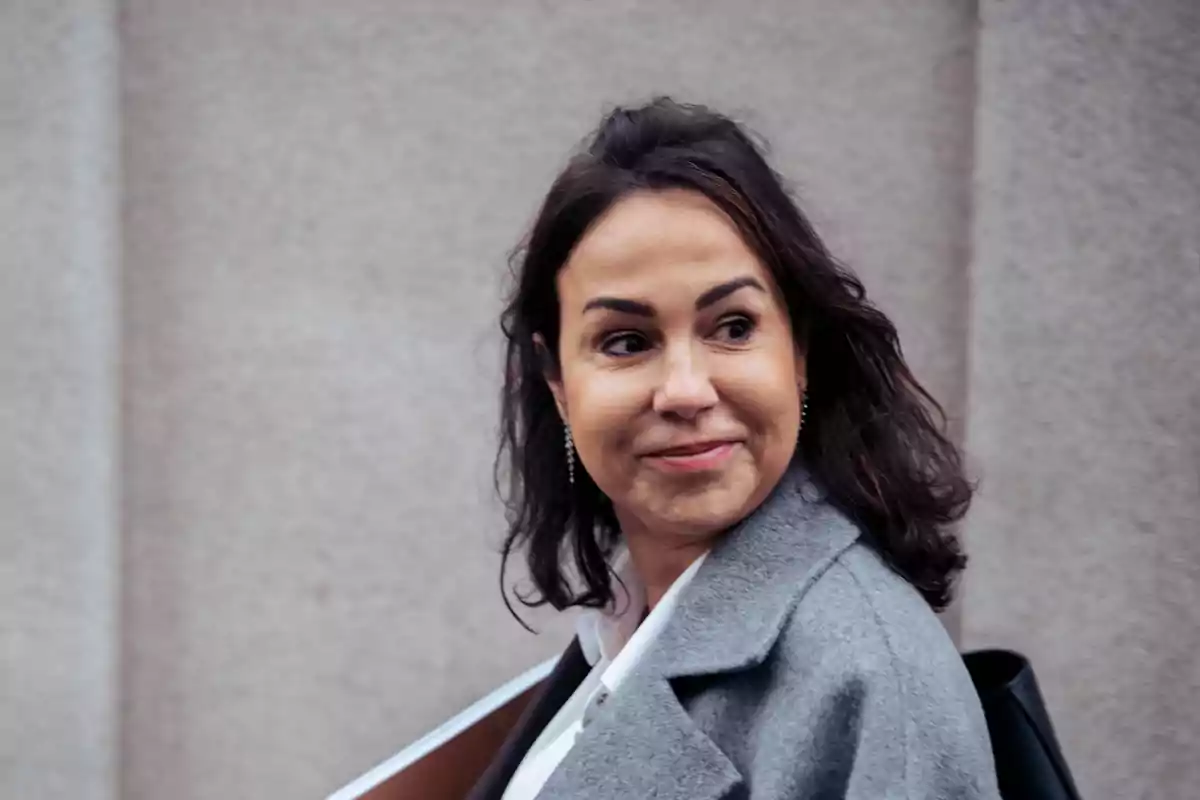 Mujer de cabello oscuro y abrigo gris sonriendo mientras mira hacia un lado frente a una pared clara Mujer de cabello oscuro y abrigo gris sonriendo mientras mira hacia un lado frente a una pared clara
