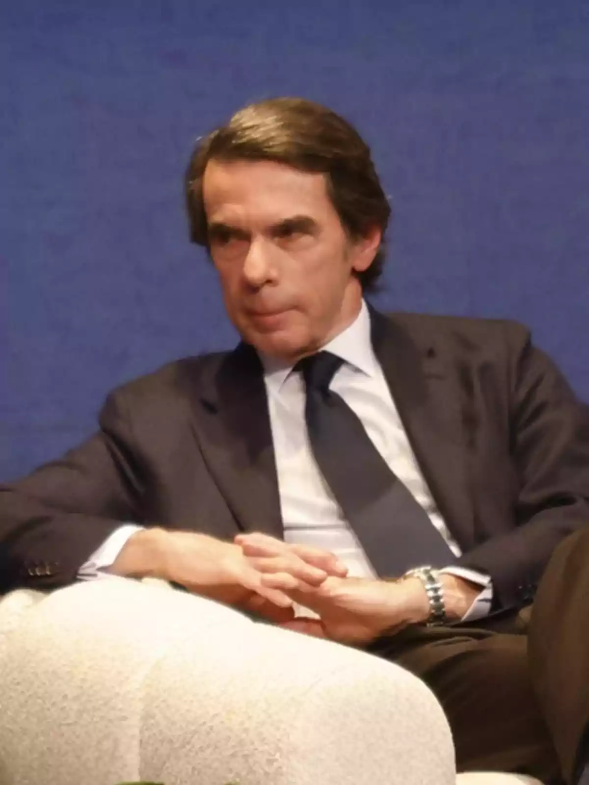 Hombre de mediana edad con traje oscuro y corbata sentado en un sillón claro sobre un fondo azul Hombre de mediana edad con traje oscuro y corbata sentado en un sillón claro sobre un fondo azul