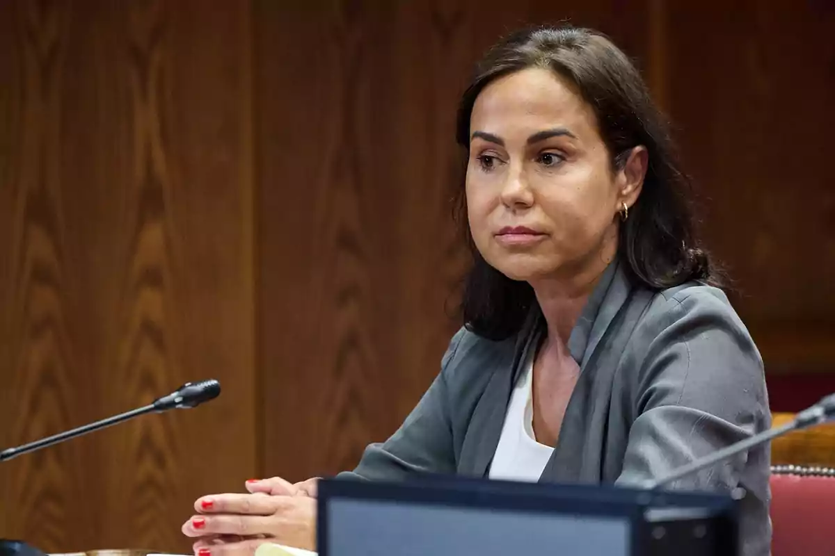 Mujer de cabello oscuro sentada en una sala de reuniones con expresión seria y las manos entrelazadas sobre la mesa