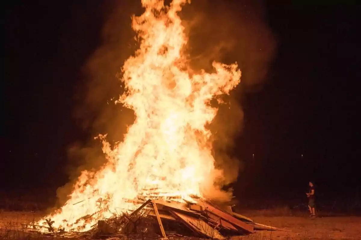 Gran hoguera ardiendo intensamente en un campo durante la noche con una persona observando a la distancia Gran hoguera ardiendo intensamente en un campo durante la noche con una persona observando a la distancia