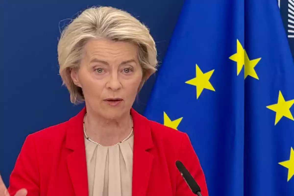 Mujer de cabello rubio con saco rojo hablando frente a un micrófono con una bandera de la Unión Europea de fondo