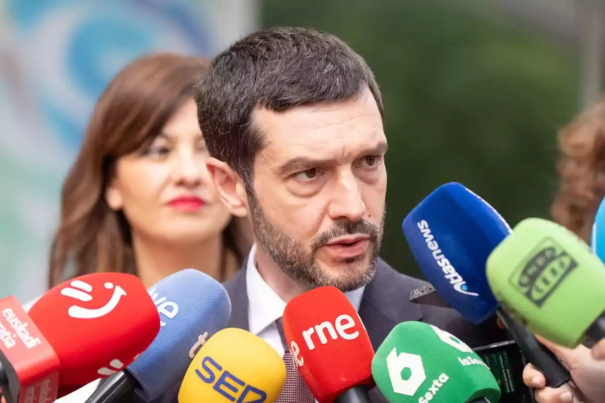 Un hombre de traje hablando ante varios micrófonos de diferentes medios de comunicación mientras una mujer lo observa en segundo plano Un hombre de traje hablando ante varios micrófonos de diferentes medios de comunicación mientras una mujer lo observa en segundo plano