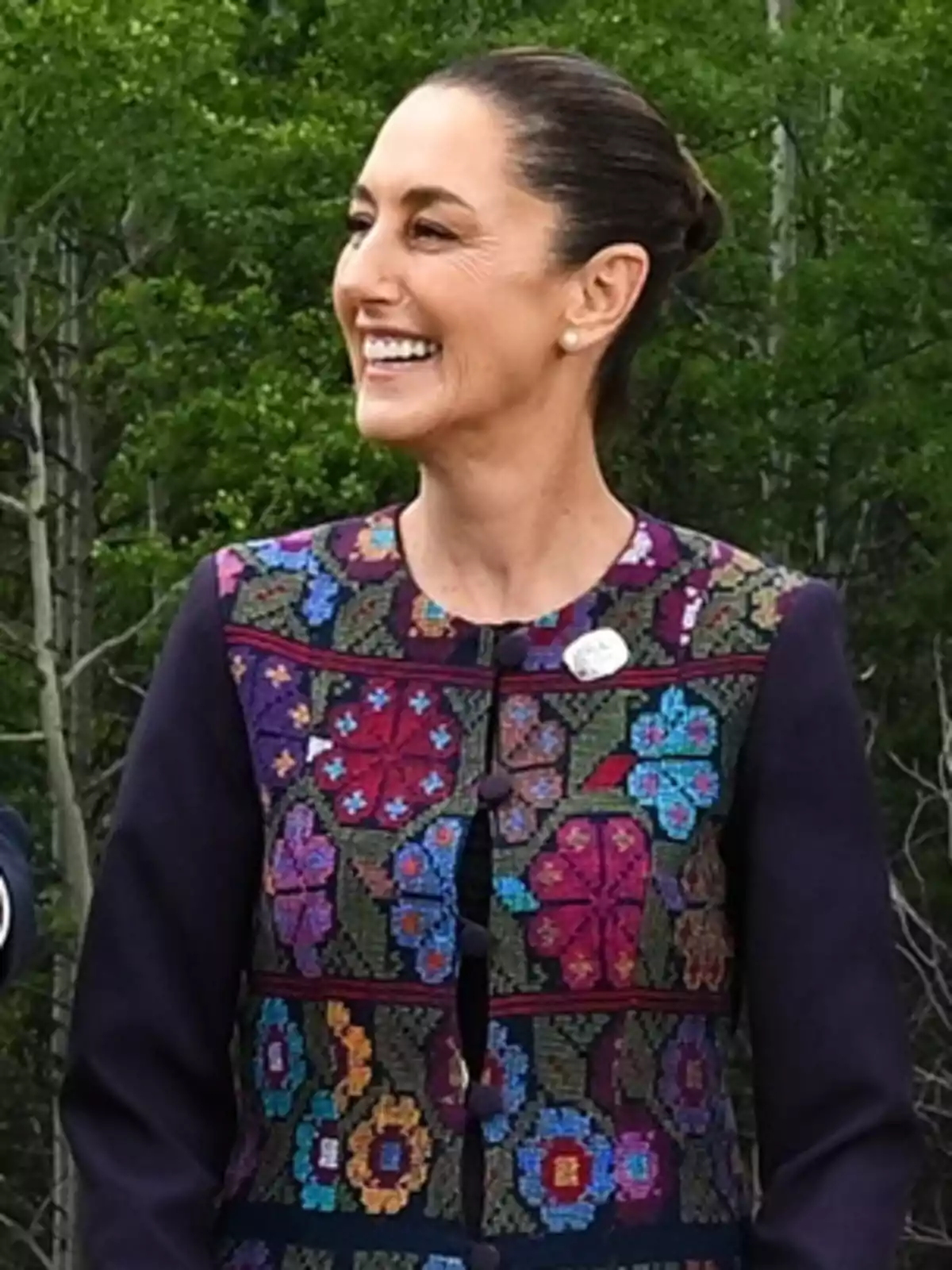 Mujer sonriente con el cabello recogido usando un saco con bordados coloridos de flores y fondo de árboles verdes Mujer sonriente con el cabello recogido usando un saco con bordados coloridos de flores y fondo de árboles verdes