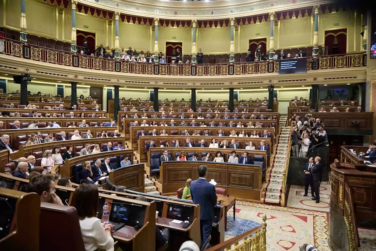 Vista general del hemiciclo de un parlamento con numerosos diputados sentados en sus escaños y una persona hablando desde la tribuna mientras fotógrafos capturan el momento