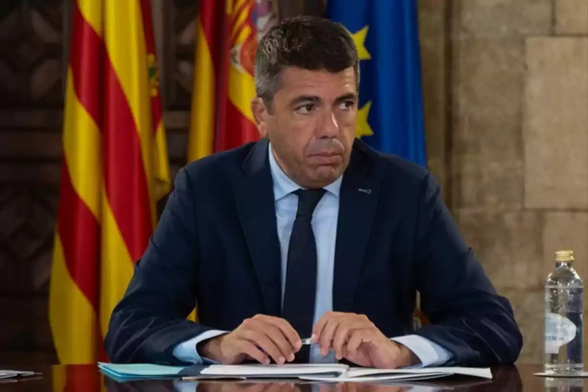 Hombre de traje azul sentado en una mesa con documentos y una botella de agua, al fondo hay banderas oficiales