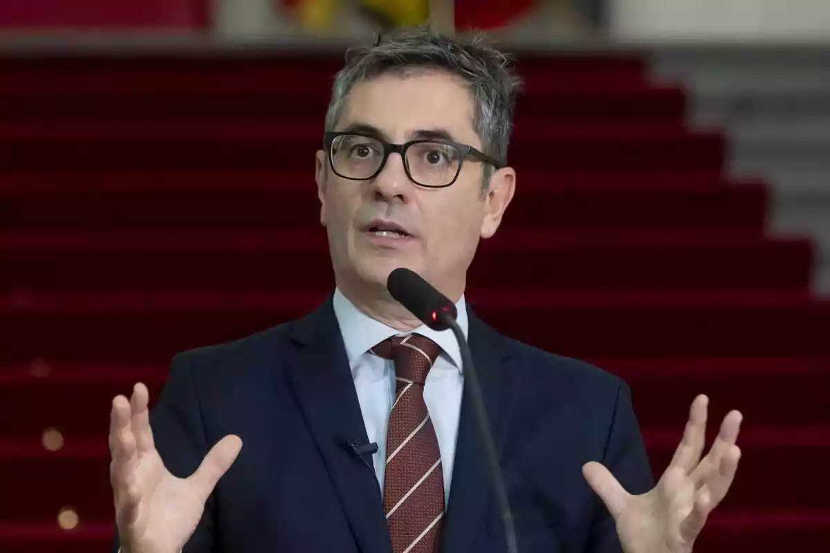 Hombre con gafas y traje hablando frente a un micrófono con fondo de escaleras rojas