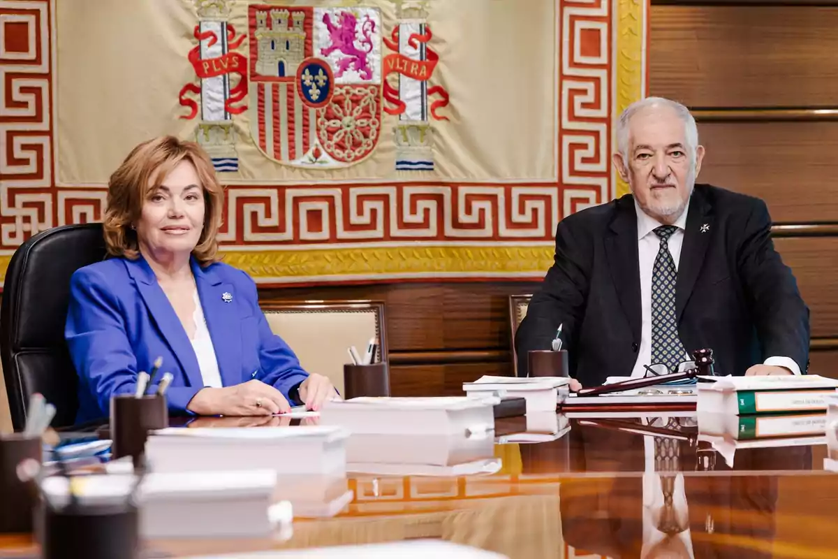 Dos personas sentadas en una mesa de reuniones con documentos y bolígrafos, al fondo una bandera con el escudo de España Dos personas sentadas en una mesa de reuniones con documentos y bolígrafos, al fondo una bandera con el escudo de España