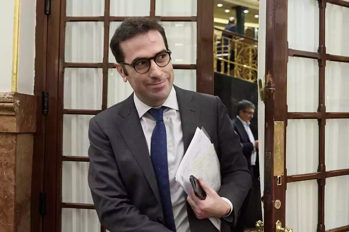 Hombre con gafas y traje sosteniendo papeles y un teléfono mientras entra por una puerta de madera en un edificio elegante Hombre con gafas y traje sosteniendo papeles y un teléfono mientras entra por una puerta de madera en un edificio elegante