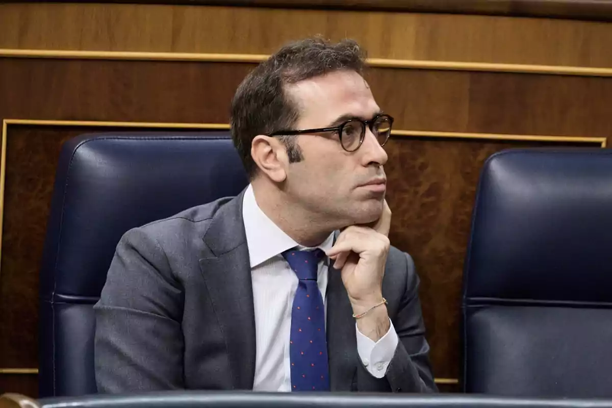 Hombre con gafas y traje sentado en una silla azul en un entorno formal mirando hacia la derecha con expresión pensativa