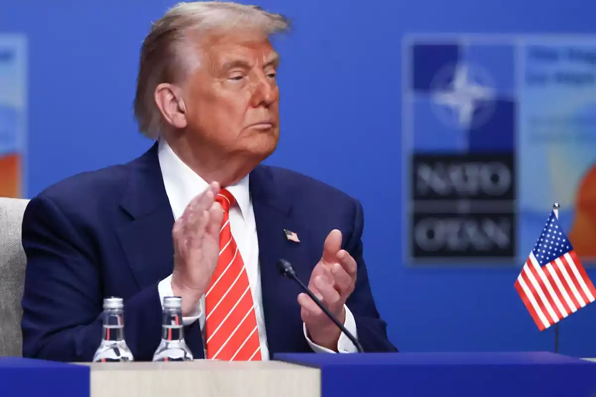 Hombre de cabello rubio con traje azul y corbata roja a rayas sentado en una mesa con una bandera de Estados Unidos y dos botellas de agua al frente fondo azul con logotipo de la OTAN Hombre de cabello rubio con traje azul y corbata roja a rayas sentado en una mesa con una bandera de Estados Unidos y dos botellas de agua al frente fondo azul con logotipo de la OTAN