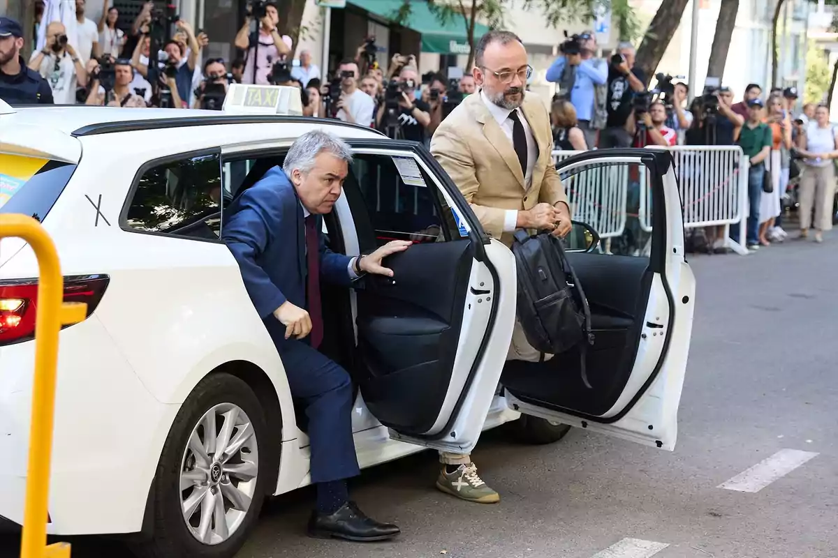 Dos hombres bajan de un taxi blanco mientras varias personas y fotógrafos los observan detrás de una valla en la calle Dos hombres bajan de un taxi blanco mientras varias personas y fotógrafos los observan detrás de una valla en la calle