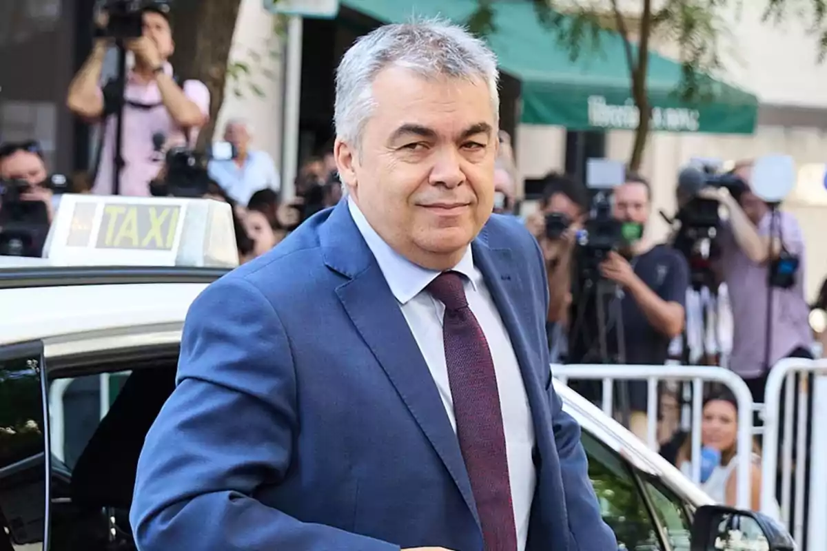 Hombre de cabello canoso con traje azul y corbata roja bajando de un taxi mientras varias personas con cámaras lo fotografían en la calle Hombre de cabello canoso con traje azul y corbata roja bajando de un taxi mientras varias personas con cámaras lo fotografían en la calle