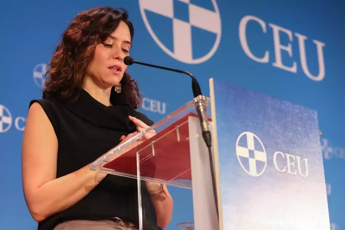 Mujer de cabello castaño hablando en un atril con micrófono frente a un fondo azul con el logo y las letras CEU.