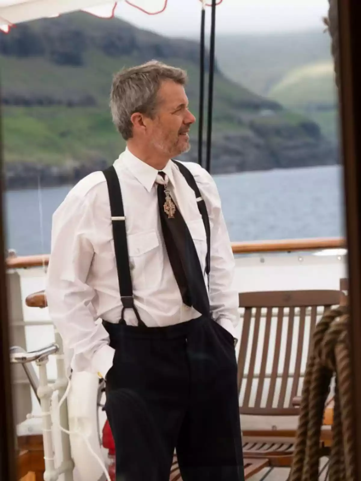 Gray-haired, bearded man dressed in a white jersey, suspenders, and a black tie, standing on the deck of a ship with a mountain and water landscape in the background. Gray-haired, bearded man dressed in a white jersey, suspenders, and a black tie, standing on the deck of a ship with a mountain and water landscape in the background.