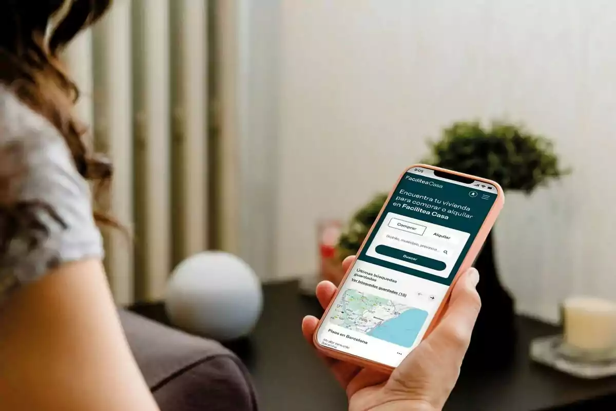 Persona sosteniendo un teléfono móvil con una aplicación de búsqueda de viviendas abierta en la pantalla, sentada en una habitación con decoración minimalista. Persona sosteniendo un teléfono móvil con una aplicación de búsqueda de viviendas abierta en la pantalla, sentada en una habitación con decoración minimalista.