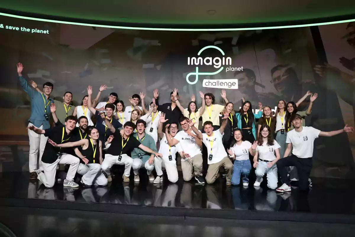 Grupo de jóvenes sonrientes posando juntos en un escenario con camisetas y credenciales, levantando los brazos frente a un fondo que dice imagin planet challenge. Grupo de jóvenes sonrientes posando juntos en un escenario con camisetas y credenciales, levantando los brazos frente a un fondo que dice imagin planet challenge.