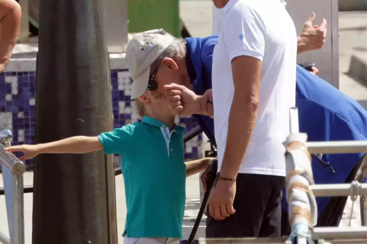 Carlitos Zurita wearing a green jersey and a cap is accompanied by two adults, one of them is King Juan Carlos I, near a railing in an outdoor setting. Carlitos Zurita wearing a green jersey and a cap is accompanied by two adults, one of them is King Juan Carlos I, near a railing in an outdoor setting.