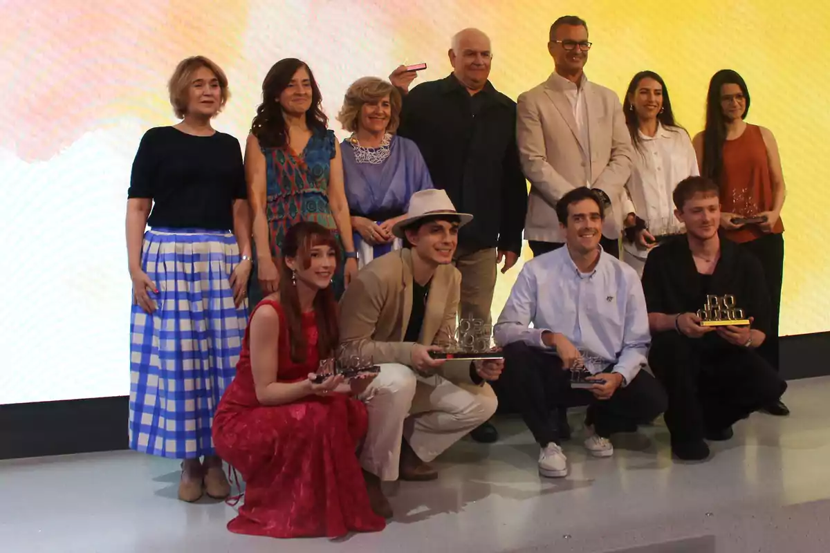 Grupo de personas posando para una foto en un evento de premiación, algunas de ellas sostienen trofeos y todos lucen sonrientes frente a un fondo colorido